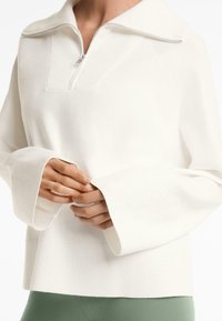 Maglia bianca con zip, colletto alto, maniche lunghe a coste e design del corpo testurizzato. Tessuto morbido con vestibilità rilassata.