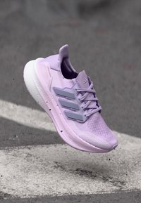 Baskets de course Adidas lavande avec semelle blanche et trois rayures grises, capturées en plein vol au-dessus d'une surface en asphalte.