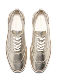 Scarpe wingtip in pelle dorata metallizzata con lacci bianchi e suole bianche, viste dall'alto, con il nome del marchio Cole Haan visibile all'interno.