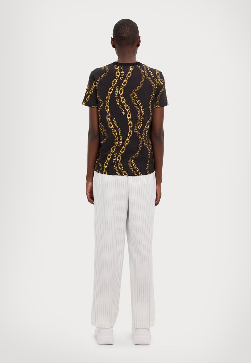 Camiseta negra con un patrón de cadenas doradas, de mangas cortas y cuello redondo, combinada con pantalones anchos blancos con pliegues y calzado blanco.