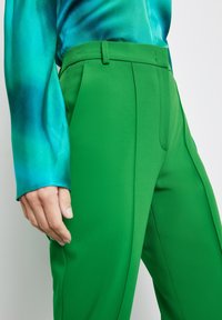 Pantalon vert vif, ajusté, avec une texture lisse et des plis sur le devant, associé à un chemisier turquoise en satin à manches longues présentant un effet dégradé.