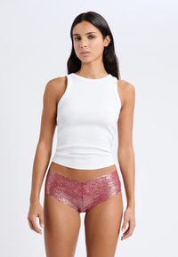 Braguitas burdeos con encaje y bordes en forma de ola, combinadas con una blusa blanca ajustada sin mangas. Se aprecian una textura suave y un delicado patrón floral.