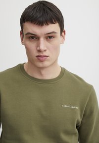 Hanorac de culoare verde măsliniu, fabricat dintr-un material moale, având un guler rotund și inscripția albă "CASUAL FRIDAY" pe pieptul stâng.
