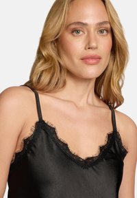 Schwarzes Camisole aus glattem Stoff mit einer gezackten Spitzenkante entlang des Halsausschnitts. Verstellbare Träger und eine lockere Passform.