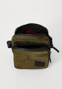 Olivengrønn nylon crossover-veske med to glidelåsrom, svart metalldeler, teksturert overflate og et svart merke på fronten.