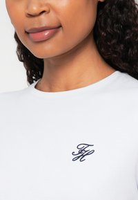 Balts kokvilnas T-krekls ar mazu, tumši zilu izšūtu logotipu kreisajā krūtīs. Apaļš kakla izgriezums un īsas piedurknes.