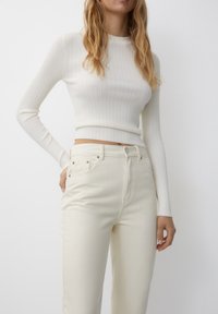 Un haut à manches longues blanc à côtes associé à un jean taille haute de couleur claire, avec un bouton et des poches, sur un fond neutre.
