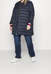 Merinovihreä toppatakki, jossa on vetoketju ja ribbikantit, punaisia ja valkoisia yksityiskohtia, yhdistettynä sinisiin farkkuihin ja valkoisiin lenkkareihin. Sujuva pinta.