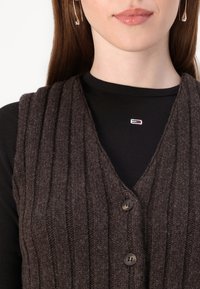 Hnedá rebrovaná vesta s hlbokým výstrihom do V, má tri gombíky a mäkkú textúru, pričom je vrstvená cez čierny top s dlhým rukávom a malým logom.