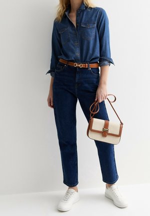 Woman wearing dark blue denim shirt and jeans with brown belt, holding beige and brown crossbody bag, wearing white sneakers.