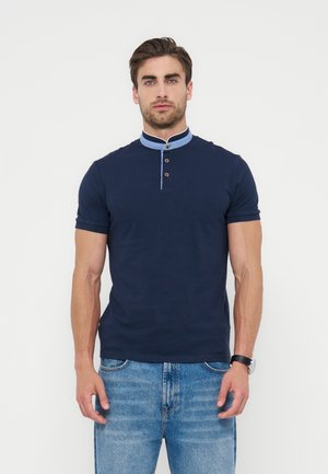 Άνδρας φοράει μπλε σκούρο κοντομάνικο πουκάμισο με ανοιχτό μπλε μανταρίνι γιακά με κουμπιά και μπλε τζιν, στέκεται μπροστά σε λευκό φόντο.