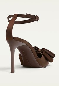 Hnědé sandály na vysokém podpatku s kotníkovým páskem a velkou dekorativní mašlí v přední části, které se pyšní elegantním stiletto podpatkem.