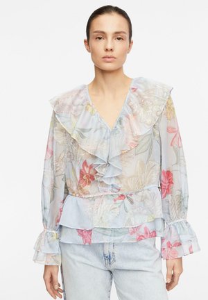 Kobieta ubrana w przezroczystą pastelową kwiatową bluzkę z marszczonym kołnierzykiem, mankietami i dołem, w połączeniu z jasnoniebieskimi dżinsami.