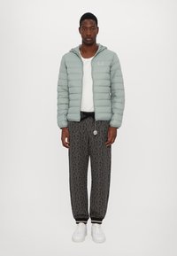 Lys grøn pufferjakke med hætte, hvid skjorte under, mønstrede sorte sweatpants og hvide sneakers. Bemærk: jakken har en logodetalje.