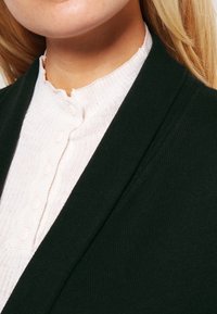 Mörkgrön cardigan i en texturerad tyg, kombinerad med en ljusrosa ribbad topp med runda knappar och hög hals.