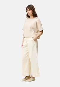 Top court beige clair avec une finition texturée, associé à un pantalon crème à jambes larges et des chaussures neutres. Le mannequin se tient avec les mains dans les poches.