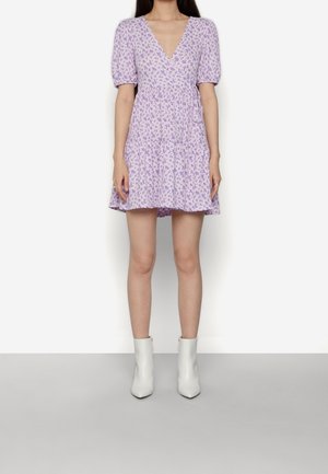 Vrouw draagt een korte lavendelkleurige bloemen wikkeljurk met pofmouwen en witte enkellaarzen, staand tegen een effen witte achtergrond.