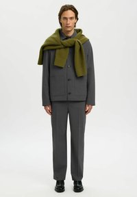 Grauer Anzug mit einer kastigen Jacke und schmal geschnittenen Hosen, verziert mit einem grünen Strickschal, der über die Schultern drapiert ist. Schwarze Schuhe runden den Look ab.