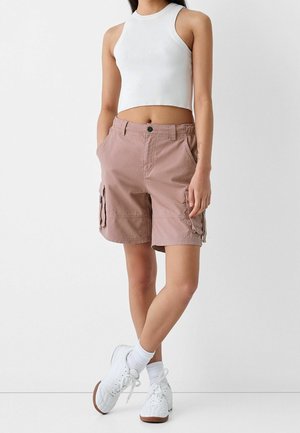 Personne portant un haut court sans manches blanc, un short cargo marron clair, des chaussettes blanches et des baskets blanches, debout devant un fond uni.