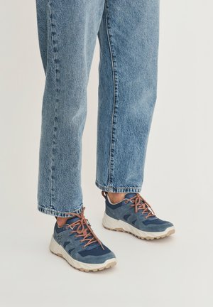 Blauwe en grijze casual sneakers met bruine veters, gedragen met lichtblauwe spijkerbroek, op een effen lichte achtergrond.