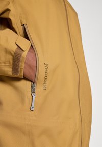 Żółta kurtka z kieszenią zapinaną na zamek z srebrną zawieszką. Logo tekstowe "X Houdini" nadrukowane na brązowo w pobliżu zamka. Gładki materiał.