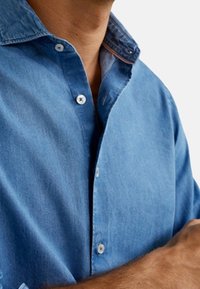 Camisa vaquera de tono azul claro, con cuello abotonado, botones blancos y un sutil detalle en contraste en el borde del cuello.