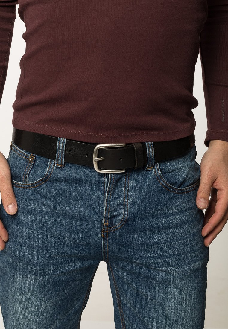 Cinturón de cuero negro con una hebilla rectangular plateada, usado sobre unos vaqueros de mezclilla azul y una camisa de manga larga color burdeos.