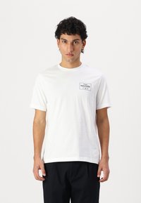 White short-sleeve T-shirt made of cotton. Features a black rectangular logo on the left chest area and a crew neckline. Clean design.
