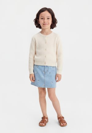 Κορίτσι με κοντά σκούρα μαλλιά που φοράει μια κρεμ ριμπ πλεκτή ζακέτα με κουμπιά, κεντημένη μπλε τζιν φούστα και καφέ σανδάλια, που στέκεται σε λευκό φόντο.