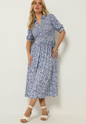 Kvinde iført en blå og hvid blomster midi kjole med rynket overdel og pufærmer, kombineret med hvide ankelrem sandaler og sølv armbånd.