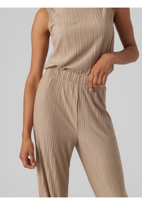 Kobieta w beżowej, prążkowanej bluzce bez rękawów i dopasowanych spodniach na gumce, pozująca z ręką opartą na talii na jasnym tle.
