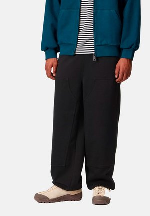 Pantaloni della tuta scuri con grandi tasche laterali, abbinati a una felpa con zip color teal e una maglietta a righe, completati con sneaker beige chiare.