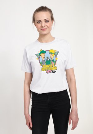 Giovane donna con maglietta bianca con stampa colorata del cartone animato Beavis & Butt-Head ’93, in piedi su uno sfondo bianco uniforme.