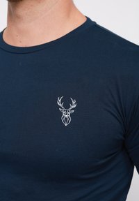 Camiseta de manga corta color azul marino con un gráfico geométrico de ciervo blanco en la parte izquierda del pecho. Fabricada con un tejido suave y liso.