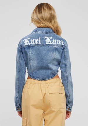 Kvinde med langt blondt hår iført en cropped blå denimjakke med teksten "Karl Kani" og beige bukser, set bagfra.