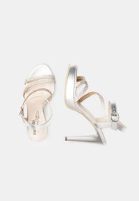 Sandali con tacco alto in argento, con tomaia metallica, design con cinturini incrociati, chiusura con fibbia e un elegante tacco a stiletto. Notevole lucentezza e texture.
