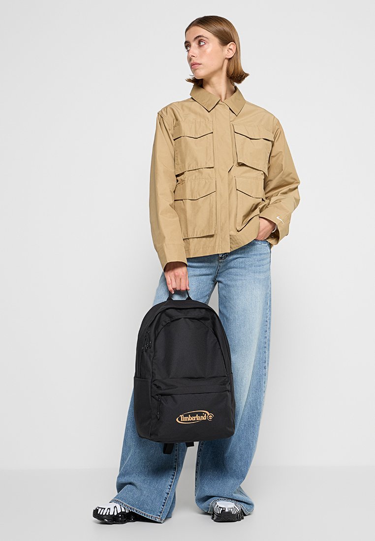 Czarny plecak z zamykanymi komorami i logo Timberland, noszony z beżową kurtką roboczą z wieloma przednimi kieszeniami oraz szerokimi dżinsami.