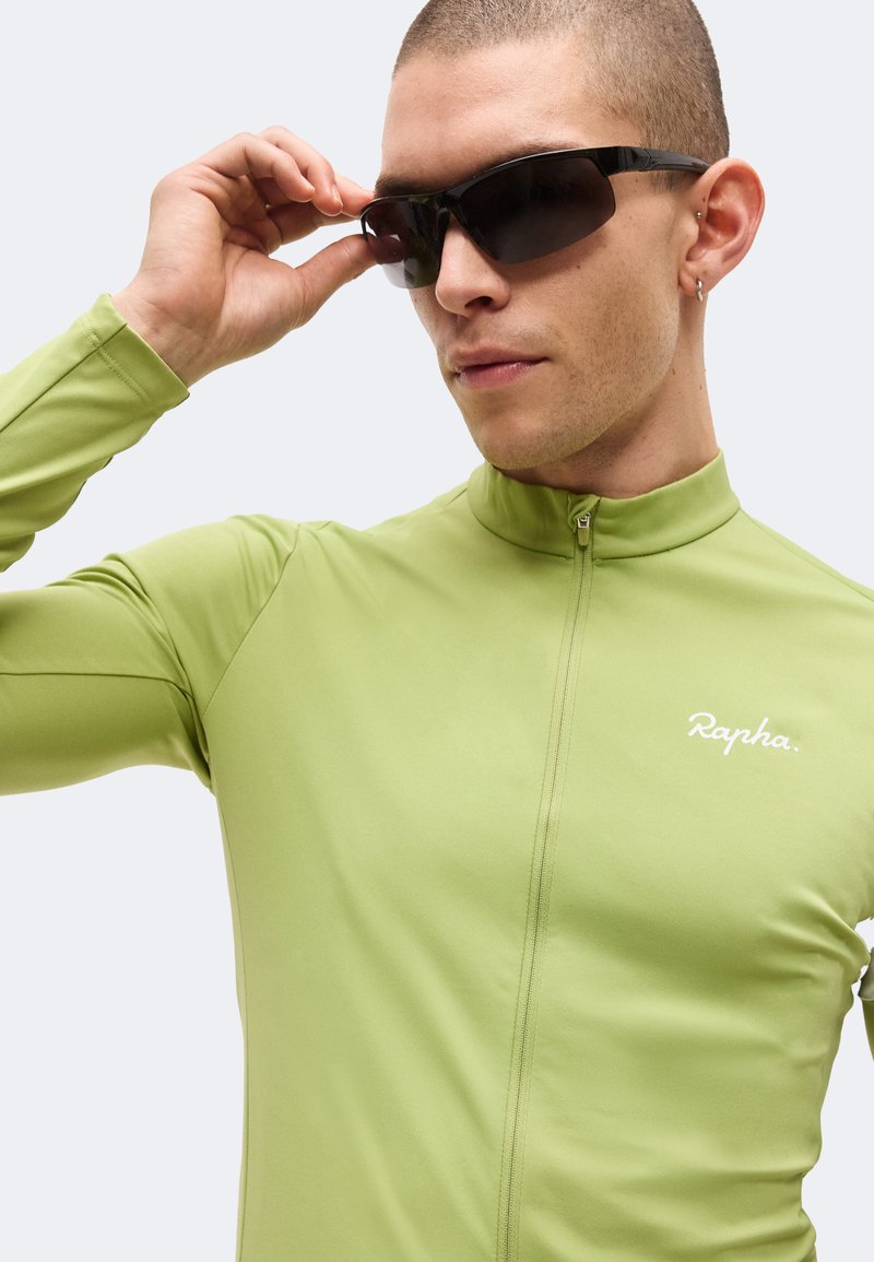 Mann trägt hellgrünes Rapha-Radtrikot und richtet schwarze Sonnenbrille vor schlichter Hintergrund.