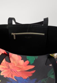 Zwarte tote bag met een bloemig patroon in levendige kleuren, voorzien van een ritsvak aan de binnenkant en een logo-detail dat op de buitenkant is gedrukt.