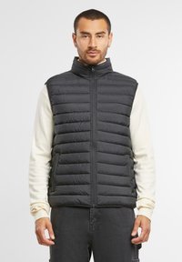 Schwarze, pufferartige, ärmellose Weste mit hohem Kragen und Frontreißverschluss. Verfügt über ein gestepptes Design und eine weiche Textur, getragen über einem cremefarbenen Langarmshirt.