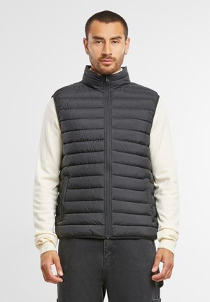 Chaleco negro, acolchado, sin mangas, con un alto cuello y cremallera frontal. Presenta un diseño acolchado y una textura suave, usado sobre una camiseta de manga larga color crema.