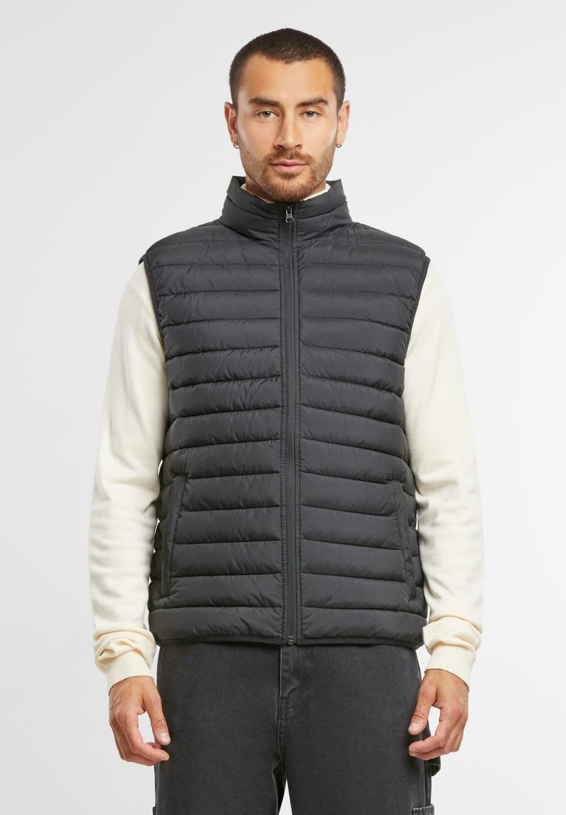 Schwarze, pufferartige, ärmellose Weste mit hohem Kragen und Frontreißverschluss. Verfügt über ein gestepptes Design und eine weiche Textur, getragen über einem cremefarbenen Langarmshirt.
