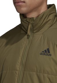 Giacca imbottita verde oliva con colletto alto, chiusura a zip e logo Adidas nero sul petto. Texture liscia e design leggero.