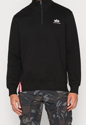 Sort halvlængde lynlåstrøje med Alpha Industries-logo på brystet, båret med grå og sort camouflage cargo bukser med flere lommer og lynlåse.