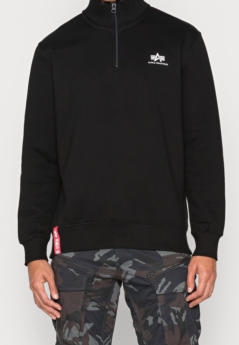 Pull noir demi-fermeture avec logo Alpha Industries sur la poitrine, porté avec un pantalon cargo camouflage gris et noir à multiples poches et zips.