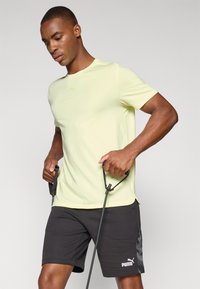 Lys gul kortærmet træningsskjorte parret med sorte shorts, der har et sidegrafiktryk og en hvid logo. Activewear-accessories holdt i hænderne.