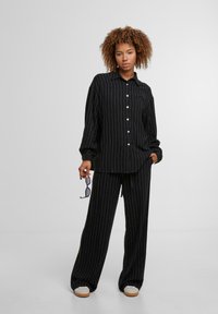 Black striped shirt with a collar and buttons paired with matching relaxed-fit pants. Worn with white sneakers and holding sunglasses.
