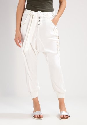 Woman wearing high-waist white jogger pants with button fly, tied fabric belt, elastic cuffs, and silver slip-on sandals.