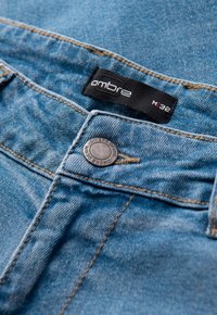 Jean en denim bleu délavé avec surpiqûres orange, doté d'un bouton en métal et d'une étiquette noire affichant la marque "ambre" et la taille M32.