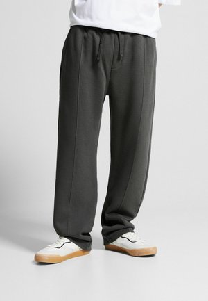 Underkrop iført løstsiddende mørkegrå joggingbukser med snørelukning i taljen og hvide sneakers med gummisåler på grå baggrund.